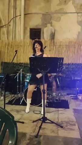 A woman with dark, curly hair and a black dress, white boots, and a necklace is singing into a microphone on a stage. She is holding a microphone in her right hand and gesturing with her left. A music stand with a black sheet of music is in front of her. Behind her, a drummer is partially visible playing a drum set. A man in a grey shirt and blue jeans is playing a guitar on the left side of the stage. There are speakers and musical equipment on the stage, with a backdrop of a bamboo-like material. The scene appears to be an outdoor performance space with a stone wall and arched doorways in the background. The lighting is dim, with some colored lights visible.