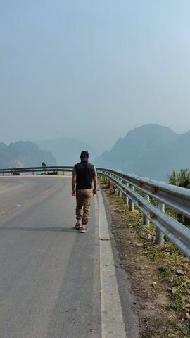 A man walks on the shoulder of an asphalt road. He wears a black vest over a blue shirt, brown pants, a baseball cap, and sneakers. He is walking away from the camera. The road is bordered by a metal guardrail. In the background are several mountains shrouded in a light haze. A motorcyclist passes him on the road, riding into the distance. The camera pans to show the view from the side of the road down into the valley. A narrow river can be seen at the bottom. The background is still hazy and the sky is overcast.