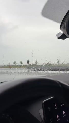 This video shows the view from inside a car looking out the windshield. The windshield is wet with raindrops, obscuring the view of the outside environment. A steering wheel is visible in the foreground. The outside environment appears to be a road with some trees in the distance. The sky is overcast and cloudy. The overall impression is of a rainy day.