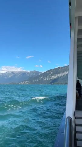 This is a first-person perspective video shot from a boat moving across a large, choppy body of turquoise water. The water is a vibrant blue-green with white foam on the crests of the waves. In the distance, a range of majestic mountains with jagged peaks and some snow-capped summits is visible under a clear blue sky with scattered white clouds. The mountains are covered in dense green trees on their lower slopes, transitioning to rocky terrain higher up. The right side of the frame shows a portion of the boat's exterior, including a white railing and some seating. A person is visible sitting on the seats at the far right of the frame. The boat appears to be moving forward, creating ripples and waves in its wake. The overall scene depicts a scenic journey across a lake or large body of water surrounded by impressive mountain scenery on a bright, sunny day.