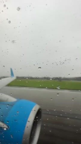 The video shows an airplane's wing and engine through a window covered in raindrops. The plane is on a runway with green grass and trees in the background. The sky is overcast. The camera is moving, suggesting the plane is either taxiing or taking off. The focus is on the details of the raindrops on the window and the engine of the plane.
