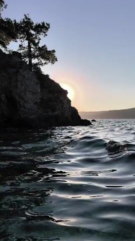 The video shows a sunlit ocean cliff with trees at the top. The camera is positioned at water level, looking towards the cliff and the sun. The sun is partially obscured by the cliff, creating a lens flare effect. The water is slightly choppy. The scene is peaceful and serene.