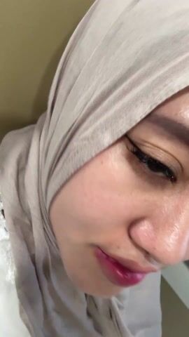 A close-up shot shows a woman wearing a light gray hijab, with a glimpse of a white, lace-trimmed garment visible. She has dark eyeliner and glossy pink lips. The camera angle is slightly tilted. Background features a plain beige wall. The woman is looking towards the camera, smiling subtly. An audio clip plays in the background featuring speech, possibly an announcement, indicating the arrival at a station.