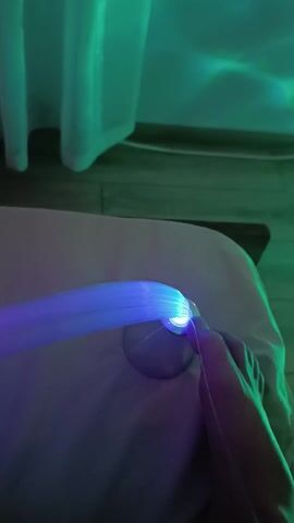 The video shows a hand interacting with a fiber optic lamp. The lamp has a round base and fibers that extend outwards. The fibers are emitting blue and green light. The hand is touching the fibers and moving them around. The background is blurred and indistinct, but there appears to be a white sheet-like object and possibly a wooden floor. The overall lighting is dim and heavily colored by the lamp's glow.