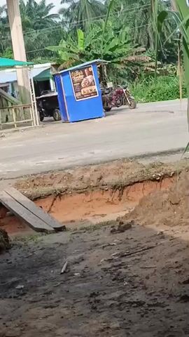 A blue food stall is set up on the side of a road. A motorcycle is parked next to it. Another motorcycle is visible further down the road. Lush green vegetation, including banana trees and palm trees, surrounds the area. A dirt excavation is in the foreground with wooden planks placed across it. The scene appears to be in a rural or semi-rural setting.