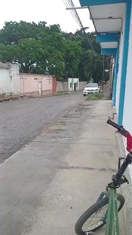 The video captures a street scene, likely in a town or village. The road is paved with asphalt and appears somewhat worn, with puddles indicating recent rain or moisture. On the right side of the street, a sidewalk runs along a building with white and blue siding. A bicycle, predominantly green with red accents, is parked on the sidewalk, leaning against something out of frame. Further down the street, a white minivan is visible, parked next to a small, light-colored structure. The background features lush green trees and other buildings, including one with a red roof and pink and white walls. Several vans are parked in a line along the left side of the street, some appearing older model and in varying colors like white, beige, and dark red. Electrical wires crisscross overhead, supported by utility poles. The sky is overcast, suggesting a cloudy day. The overall atmosphere is quiet and still.