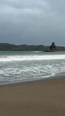 The video shows a beach scene. The sand is dark and wet near the water's edge, transitioning to a lighter hue further inland. Waves, some breaking with noticeable force, roll towards the shore. The water is a greenish-gray color. In the background, a rocky outcrop or stack rises from the water, and behind that are rolling green hills under a heavy, overcast sky.