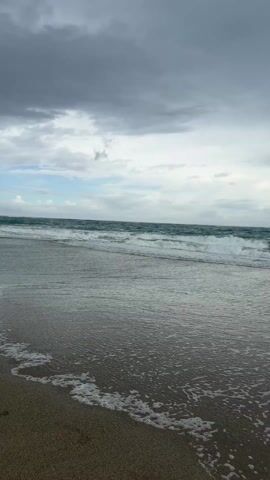 The scene is a beach on a cloudy day. The sky is overcast with gray and white clouds. The ocean water is a dark turquoise color with small waves rolling in towards the shore. The sand is dark and wet, with a thin layer of foamy water washing over it. There are no people or other objects visible in the frame. The camera is stationary, capturing the natural scene.