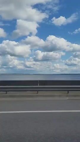The video shows a view from a moving vehicle, likely a car or truck, traveling alongside a large body of water under a blue sky with white clouds. A gray asphalt road is in the foreground, with a white line marking the lane. A metal guardrail separates the road from a concrete barrier, beyond which lies the expanse of the water. The water appears calm and stretches to the horizon. In the background, land is faintly visible across the water, suggesting a distant shoreline. A black SUV passes the camera in the middle distance, moving in the same direction. The scene suggests a daytime drive on a clear day.