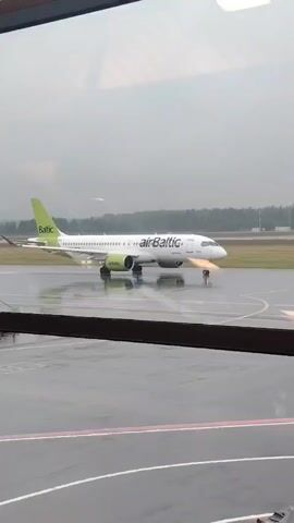 A video shows an Air Baltic plane on an airport runway, seen from inside an airport terminal through a window. The plane, painted white with green and black accents, has the "airBaltic" logo on its fuselage and "Baltic" on its tail. The runway is wet, indicating rain or recent rain, and reflections are visible on its surface. Trees and what appears to be part of an airport building are visible in the distant background. The camera pans slightly to the right, following the plane as it slowly moves. The video is shaky at times.
