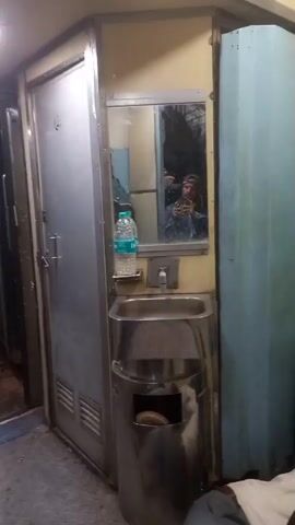 The video shows the interior of a train bathroom. A small stainless steel sink with a tap is positioned below a mirror. A bottle of water rests on a soap dish beside the mirror. The mirror reflects a man holding a cell phone, presumably recording the video. He wears a blue shirt and a baseball cap. The bathroom has a metal door with ventilation slats and a blue partition wall. The floor has a diamond pattern. The train is in motion.