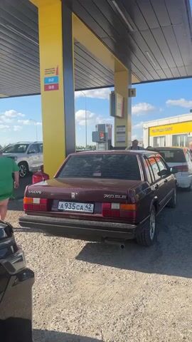The video shows a gas station with several cars. A burgundy Volvo sedan is parked at a gas pump. A woman in a green dress is walking away from the Volvo. Another car, a white SUV, is parked behind the Volvo. In the foreground, part of a black car is visible. The gas station has yellow pillars and a black roof. There are signs with numbers indicating gasoline prices. The sky is blue with some clouds. The ground is covered in gravel. The video pans to show the interior of a black minivan, then the exterior of the minivan. A white car is parked next to it.