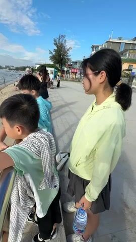 A group of four children are standing on a paved walkway overlooking a beach. The child closest to the camera, a boy wearing a green t-shirt with a plaid cloth draped over his shoulder, is leaning on a metal railing. To his left, another boy in a blue shirt also leans on the railing. Further back, a third boy in a light blue shirt and a young girl in a yellow jacket with glasses and her hair in a ponytail stand. The girl is holding a bottle of water. On the beach, several people are visible, some are in the water, some are sitting on the sand, and one person is digging in the sand. Beach umbrellas are scattered on the sand. The sky is blue with white clouds. Mountains are visible in the distance across the water.
