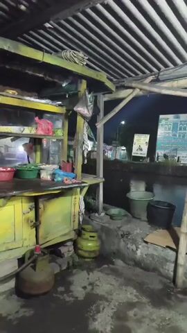 This media file captures a street food stall in Sidorejo, Indonesia, at night. The stall is a simple, open-air structure with a corrugated metal roof supported by wooden beams. A bright fluorescent light illuminates the workspace.

The main subject is a person, seen from the side, wearing a green t-shirt and dark pants, standing in front of a cooking setup. They appear to be operating a food stall, with a large wok over a gas burner. The wok is smoking, suggesting active cooking. Nearby, there are various kitchen items, including large green buckets, a smaller green bowl, and what appear to be propane gas cylinders. The stall itself is constructed with weathered wood and metal, painted a vibrant but peeling yellow. Shelves above the counter hold red and green bowls, and some plastic bags.

The setting is a street at night. The ground is wet and appears to be concrete, with puddles reflecting the artificial light. In the background, a street is visible with passing vehicles, indicated by their headlights and taillights. Further back, there are buildings and signs, one of which is a large blue and white poster with text in Indonesian. The overall atmosphere is bustling and typical of a nighttime street food scene in a developing urban area. The presence of the gas stove and wok points to food preparation, likely for sale to passersby. The lighting suggests it's after dusk, and the wet ground might indicate recent rain.
