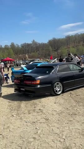 The video shows a gathering of cars and people in a dirt lot. Several cars, including black, teal, white, and red models, are visible, many with custom wheels. People are standing around, talking, and observing the cars. The environment includes trees, a dirt hill, tents, and power lines against a partly cloudy sky. The camera moves, showing the vehicles and people from different angles. The overall scene depicts a car show or meetup.
