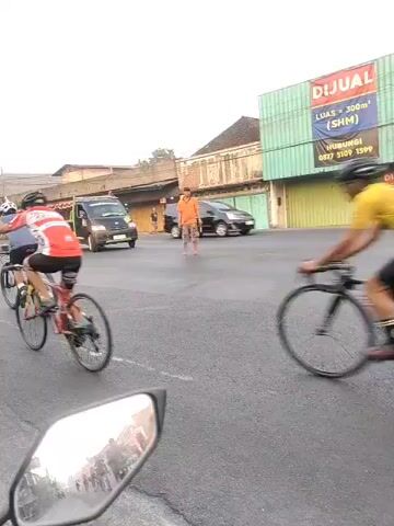 This is a video taken from a moving vehicle, likely a motorcycle, showing a group of cyclists riding on a paved road. The cyclists are wearing helmets and cycling attire. Some vehicles, including a small truck and a scooter, are also present on the road, suggesting a public street. The environment appears to be an urban or semi-urban area with buildings visible in the background. The video captures the movement of the cyclists and the surrounding traffic.