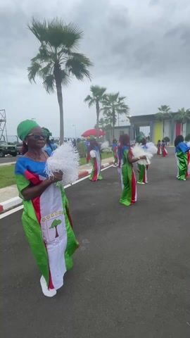 A group of approximately eight women are captured participating in an outdoor performance or ceremony on a paved road. They are dressed in vibrant, flowing garments that incorporate the national colors of Equatorial Guinea: green, white, and red, with blue accents on the top. Each woman holds white, feathery pom-poms or fans, and some are gently swaying or moving. The woman in the foreground wears a green headscarf and glasses, facing towards the right. The dresses feature an emblem on the white stripe, which appears to be the coat of arms of Equatorial Guinea, including a tree design.

In the background, several tall palm trees line the road, indicative of a tropical climate. Modern buildings are visible further in the distance under an overcast sky, suggesting daytime with cloudy weather. The scene appears to be a formal outdoor setting, possibly an entrance or public area in Malabo, Equatorial Guinea, given the national attire and location context.