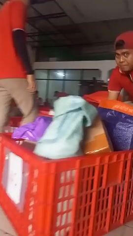 The video shows an indoor scene, likely a warehouse or distribution center. Several people, some wearing red shirts and caps, are visible. They appear to be involved in sorting or packing goods. In the foreground, a red plastic crate is filled with packages and bags. One person is seen pushing the crate. In the background, other individuals are seated on the floor, also working with packages. The environment includes tiled flooring, white walls, and industrial fans mounted on the ceiling. The lighting is somewhat dim, with fluorescent lights visible. The overall impression is of a busy workplace focused on logistics and package handling.