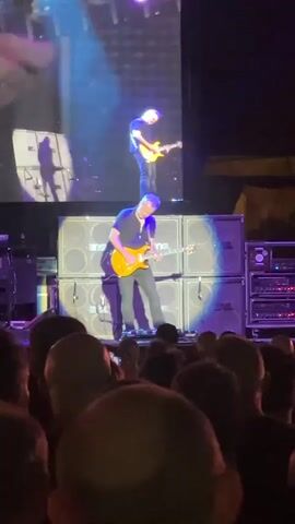 A concert crowd is visible in the foreground, with their heads and shoulders filling the bottom of the frame. In the mid-ground, a man stands on a stage, playing a yellow electric guitar. He is wearing a dark t-shirt and grey pants. Behind him, a stack of large, square speaker cabinets with circular grills are visible. Above this, a large screen displays an image of another man playing a yellow electric guitar, silhouetted against a bright light. The stage is lit with blue and white spotlights. The overall environment suggests an indoor concert venue.