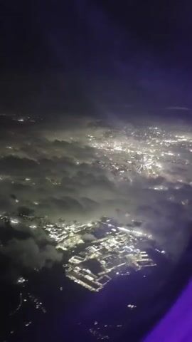 The video shows an aerial view of a city at night, taken from an airplane window. The city lights are visible through a layer of clouds. The perspective suggests the plane is descending or flying low over the urban area. The purple edge of the airplane window is visible in the lower right corner.