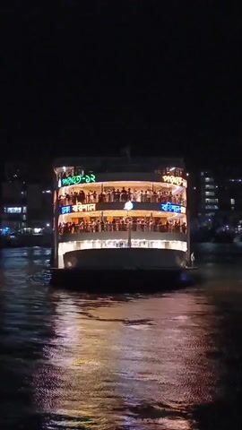 The video shows a multi-story ferry boat illuminated at night, moving on a dark body of water. The boat is filled with people visible on its open decks.  Buildings and other city lights are visible in the background. The water reflects the lights from the boat.