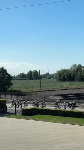 A group of cyclists are riding in a line formation on a paved track under a clear blue sky. The cyclists are wearing helmets and athletic clothing. They are moving from left to right across the frame. In the background, there is a grassy area, a fence, agricultural fields, trees, and some buildings. The sun is shining, casting shadows.