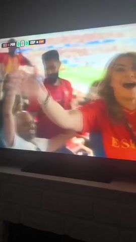 The video shows a television screen displaying a soccer match. In the foreground, a woman in a red shirt with "ESPANA" written on it is cheering and waving. The soccer match itself is being played on a green field with white lines. The scoreboard shows the score as 0-0. There are spectators in the background wearing red and white.