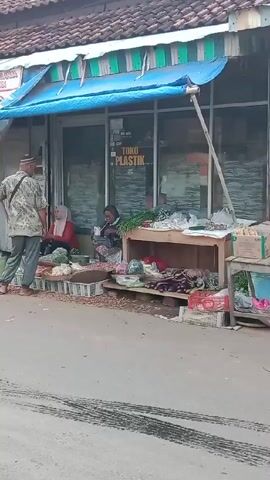 A man walks up to a small roadside stall selling vegetables. Two women are sitting behind the displayed produce. One woman is wearing a red hijab, the other is wearing a headscarf. The sign above the stall says "Toko Plastik", indicating it may sell plastic goods in addition to vegetables. The man speaks to the women. The stall is set up in front of a building with a turquoise awning. The road is asphalt with tire tracks.