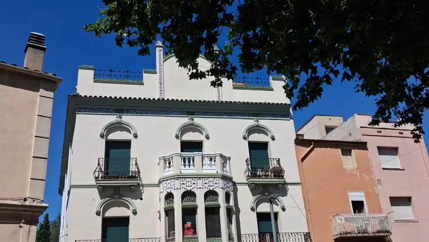 The video shows a three-story building with a white facade. The building has decorative elements, including a row of windows with small balconies and green shutters on the first and second floors. The ground floor features three arched entrances with closed green doors. The building is situated on a paved square with planters and some black posts with names on them. To the right of the white building is a peach-colored building, and to the left is a beige building.  The sky is blue and sunny.  A red Elmo toy is visible in one of the windows on the second floor. The camera slowly tilts down, showing more of the square. A tree branch partially obscures the top of the white building.