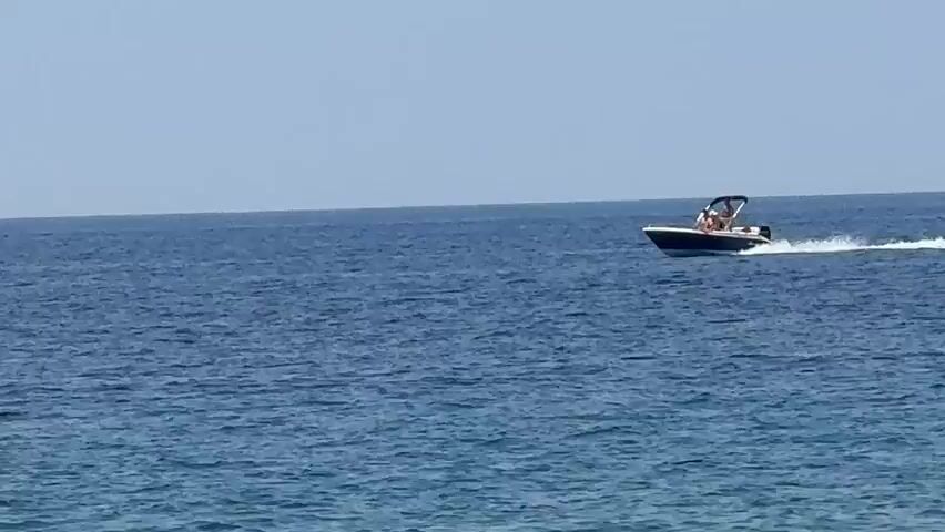 A small speedboat with a black canopy is moving from left to right across the frame, creating a white wake behind it. There are at least three people visible on the boat, seated. The boat is on a large body of blue water under a clear, pale blue sky. The water appears calm with small ripples.
