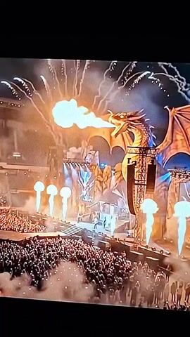 The video opens with a wide shot of a stadium or arena with a large stage elaborate stage set that appears to be designed as a fantasy castle with a prominent dragon prop. The dragon is breathing fire, and there are pyrotechnics and smoke effects. A large crowd of people is visible in the stands. The scene then cuts to a close-up of a bald man with a beard and sunglasses, wearing a leather jacket and singing into a vintage microphone. The lighting is red and dramatic, giving a close-up view of the man's face as he sings intensely.