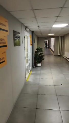 The video shows a hallway with a gray wall with three yellow signs with black arrows pointing left. The signs have words written on them in a language that is not English. On the wall is a picture of a large yellow mining truck against a rocky mountain backdrop. The hallway has gray floors and a door that leads to another area with a plant.