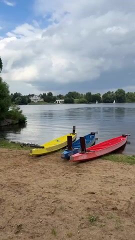 The video shows a side view of three dragon boats lined up on a sandy shore next to a lake. The boats are colored yellow, blue, and red, and they are facing the water. The lake is calm, with some ripples on the surface. In the background, there are trees and buildings on the opposite shore, and a cloudy sky with some patches of blue. There are no people visible in the video. The video appears to be a static shot with no camera movement.