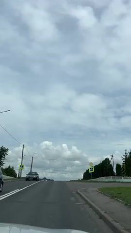 A vehicle drives down a road. The road is paved and has white lane markings. There are trees and grassy fields on either side of the road. The sky is cloudy. There are traffic signs and streetlights visible. In the distance, there is a slight uphill slope to the road.