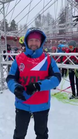 A man in a blue and red vest with the word “судья” (judge) written on it is flexing his arms and smiling at the camera while standing in a snowy outdoor setting. He is wearing a blue hooded jacket, black pants, gloves, and a pink hat. In the background, there are other people wearing helmets and ropes, suggesting a climbing or obstacle course event. A metal structure is visible behind the man, along with some trees. The snow-covered ground and the overcast sky indicate a cold, winter day. The man is the main focus of the video and appears to be in good spirits, possibly related to the event he is participating in.
