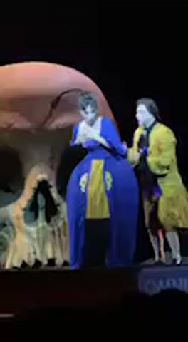 The video captures a theatrical performance on a dark stage. Two performers are visible, dressed in period costumes. One wears a prominent blue dress with yellow accents, and the other a yellow coat over white trousers. They are interacting dramatically, appearing to struggle or engage in a chase around a very large, light-colored skull prop that dominates the left side of the stage.

Initially, the performers are illuminated in front of the skull. The person in blue seems to be pulled or led by the person in yellow. As they move, the stage lighting shifts, becoming darker with blue and purple hues, particularly as the performers move behind the large skull prop and partially disappear from view. Another smaller, darker skull prop is briefly visible on the far left. Opera or theatrical singing is audible throughout the clip. Part of the word "OMNIA" is visible on a stage element in the bottom right corner.