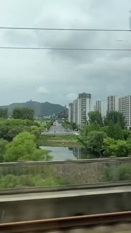 This video captures a dynamic scene from the perspective of a moving train in Gongshu District, China. The primary subjects are the urban landscape and natural scenery blurring past the window. We see a dense expanse of vibrant green trees dominating the middle ground, suggesting a temperate climate and lush vegetation. Beyond the trees, a cluster of modern, tall white buildings with visible windows are arranged in a line, indicative of a developed urban area. In the background, a large, verdant mountain range rises, its slopes covered in trees. A noticeable industrial structure, resembling a tall tower with scaffolding, stands on the mountain. The sky is overcast with heavy, grey clouds, hinting at a cloudy or potentially rainy day. The time of day is difficult to ascertain precisely due to the cloud cover, but it appears to be daytime. The blurred effect of the moving train is evident, especially in the foreground where the train tracks are visible and the scenery appears streaky. There is no visible text in the video. The overall impression is one of transit through a region that blends natural beauty with modern development.