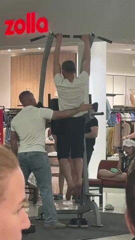 A man in a white t-shirt and black shorts is performing pull-ups on a pull-up bar. Another man in a white t-shirt and blue jeans is standing next to him, with his hands on the pull-up performer's back and waist, assisting him. The pull-up bar is part of a larger metal structure, possibly a home gym or workout station. In the background, there is a clothing store called "zolla" with racks of clothes visible. Several other people are in the background, some looking at the men, and one person is seated on a bench. The environment appears to be indoors, possibly a shopping mall or a large retail space.