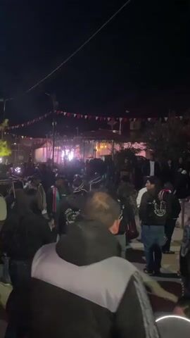 A crowd of people gathers outside at night. The Chilean flag flies next to a church with a religious figure decorated with red and white flowers on its roof. Men wear black jackets with the words "LAKITAS DE LIMAXINA MALCONES" on the back. The crowd is watching or participating in an event with loud music. Some are standing, while others are moving around. The event space is decorated with triangular flags. A light pole illuminates the crowd.