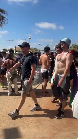 The video shows a crowded outdoor scene under a bright blue sky with fluffy white clouds. The ground is paved with reddish-brown material. Several palm trees are visible, with one tall building in the background on the right. A line of people is moving towards the right of the frame. The people are casually dressed, many in summer attire such as shorts, tank tops, and swimwear. Some individuals are carrying rainbow-colored items, suggesting a pride event. A man in the foreground on the right is wearing a light yellow halter top and white pants. To his left, a man in a white t-shirt and black shorts is walking away from the camera. Further left, a man in a blue bucket hat and gray shorts is walking towards the camera, carrying a rainbow flag draped over his shoulder. He has a shopping cart beside him, filled with various items. In the background, several people are visible, including a shirtless man and two individuals in what appear to be security uniforms. One of the security personnel is facing away from the camera, and the other is partially visible from the side. The overall atmosphere appears to be festive and social.