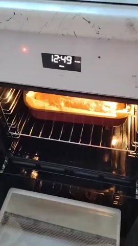 The video shows a close-up view of a rectangular cake or bread baking inside an oven. The cake is in a reddish-brown baking pan lined with parchment paper. The top of the baked good is golden brown and appears to be slightly risen and bubbling in some areas. The oven racks are visible below the baking pan, and the interior of the oven is illuminated by a warm light. The oven's digital display shows the time as 12:49. The shot is slightly angled, looking into the oven from the front.