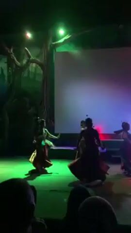 The video depicts a dance performance taking place on a brightly lit stage. Five dancers wearing long, dark gowns and headpieces are performing against a backdrop of a large white screen illuminated with changing colors (red, green, blue, purple). The stage itself is painted green and features a decorative tree prop on the left side. The lighting is dynamic, with spotlights moving across the stage and changing colors. There is a small audience present, visible in the foreground.
