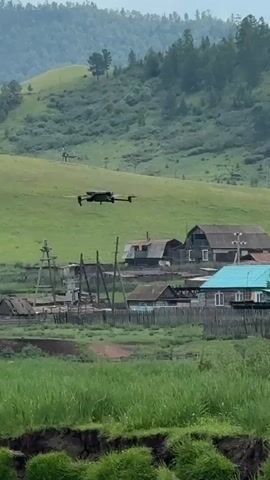 A drone is captured mid-flight over a rural village. The drone, a quadcopter with a dark body and visible propellers, is positioned in the upper left quadrant of the frame. It appears to be moving from left to right. Below the drone, a cluster of houses with varied roof colors, including turquoise and brown, forms the village. Wooden fences delineate the properties. The landscape behind the village features rolling green hills dotted with trees. The sky is overcast. The overall scene depicts a peaceful, rural setting.