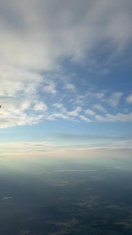 The video shows a view from an airplane window as it flies over a landscape. The plane's wing, with a red logo, is visible in the foreground on the left side of the frame. Below the wing, the ground appears as a dark expanse with patches of lighter colors, suggesting fields or settlements. Rays of sunlight break through the clouds, illuminating parts of the landscape. The sky above is a mix of blue and white clouds, with the sun positioned towards the horizon, creating a soft, hazy light. The overall scene depicts a peaceful, aerial view during daylight hours.