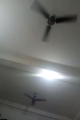This is a shaky, blurry video taken from below, looking up at a ceiling. Two ceiling fans are visible, both in motion, with their blades blurred by the speed. The ceiling is white, with a light fixture in the center of the frame emitting a bright, hazy light that washes out some of the detail. The fans are dark in color, and one is larger and closer to the viewer than the other. The overall impression is of a room with ceiling fans running.