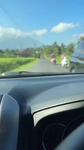 The video shows a view from inside a vehicle, looking out through the windshield at a rural road. The road stretches into the distance, lined with lush green fields on either side. In the background, a mountain is visible under a clear blue sky with scattered clouds. Several motorbikes are seen on the road, with people riding them. The overall scene depicts a scenic drive through a countryside landscape.