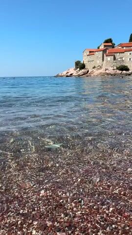 The video showcases a serene coastal scene at Sveti Stefan, Montenegro. The foreground is dominated by a clear, shallow body of water lapping over a pebble beach. The pebbles are a mix of earthy tones, with reddish-brown hues being particularly prominent. The water is so transparent that the pebbles on the seabed are clearly visible, creating a mesmerizing pattern as the light refracts.

In the mid-ground, the gentle waves of the Adriatic Sea create a shimmering surface. A light-colored object, possibly a piece of debris or a larger sea creature, drifts through the water. The sea stretches out towards the horizon, meeting a clear, bright blue sky.

The background features the iconic Sveti Stefan island, a small fortified islet connected to the mainland by a narrow causeway. The island is adorned with several ancient stone buildings, their terracotta roofs standing out against the lush green of the trees and the blue of the sky. The buildings are clustered on a rocky outcrop, adding to the picturesque and historic ambiance of the location.

The time of day appears to be late morning or early afternoon, indicated by the bright sunlight casting clear shadows and the intensity of the blue sky, suggesting fair weather. The overall mood conveyed is one of tranquility and natural beauty, inviting viewers to imagine a peaceful day by the sea. There is no visible text in the media file.