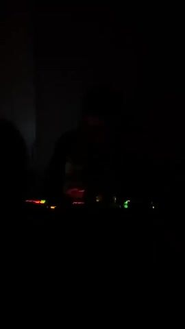 The video is recorded in very low light and shows a person standing at a DJ station.  Colored lights are visible on the station. The person fades from view as the video progresses. The environment appears to be a dark room.