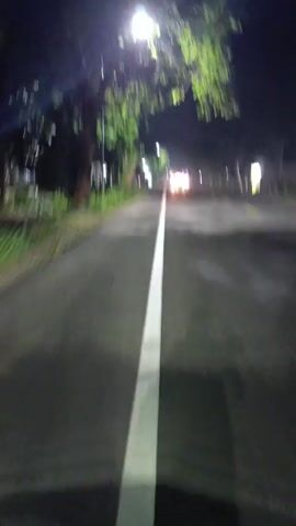 The video is shot from the perspective of a person riding a motorcycle at night. The road is paved and marked with a solid white center line. There are trees and buildings visible along the roadside, illuminated by streetlights. Several vehicles with headlights are approaching in the distance. The camera moves forward, following the road. The shadow of the motorcycle rider is visible on the road in front. The sound of wind is audible, indicating the speed of the motorcycle.