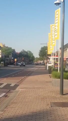 A person is walking in front of a Jumbo supermarket. The video pans from left to right, showing the exterior of the supermarket and the street in front of it. The supermarket has a black facade with a large yellow "JUMBO" sign. There are flags with the Jumbo logo on poles outside the store. The street is paved with bricks and has a sidewalk. There are cars driving on the street. The sky is clear and blue.